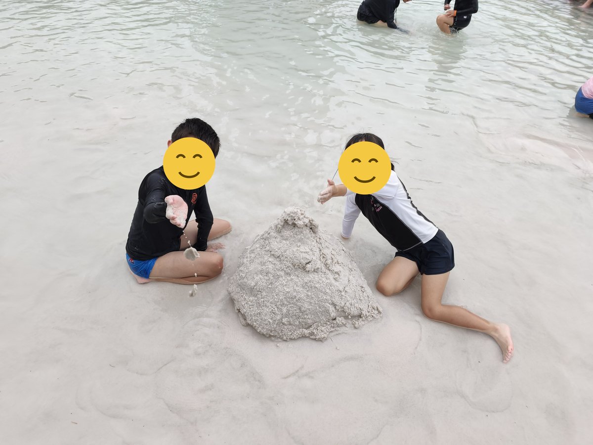 屋外プールの浅瀬で砂の山を作る子ども2人