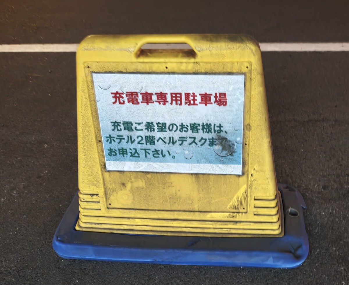 ホテル駐車場に置かれた黄色い看板。充電車専用駐車場、充電ご希望のお客様はホテル2階ベルデスクまでお申込下さいと書かれている